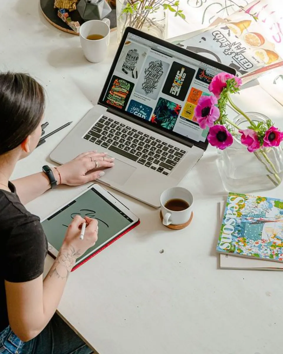 A woman working on a laptop at a bright desk with coffee and a tablet